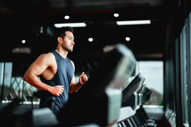 Person running on a treadmill in a well-lit gym, wearing a sleeveless dark blue shirt and appearing focused. Multiple treadmills are lined up, with large windows in the background letting in natural light, creating a bright and modern workout environment.