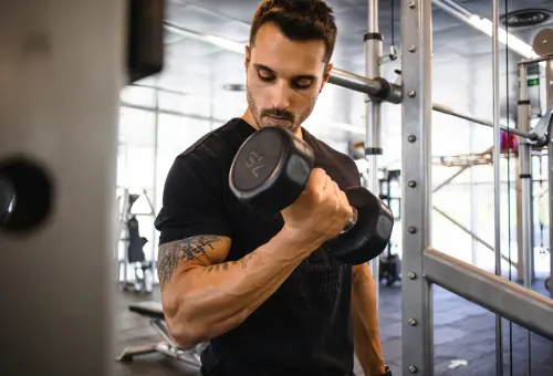  A person wearing black t-shirt performs a bicep curl with a 17.5 kg dumbbell in a naturally lit gym, emphasizing focused strength training and muscle activation.