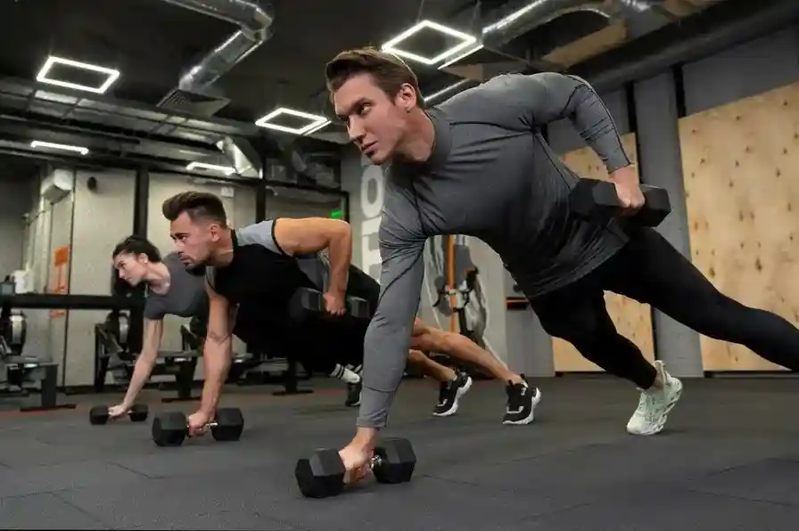 Three individuals hold plank positions with dumbbells in a sleek gym, demonstrating balance, core strength, and coordination during a strength-focused workout.