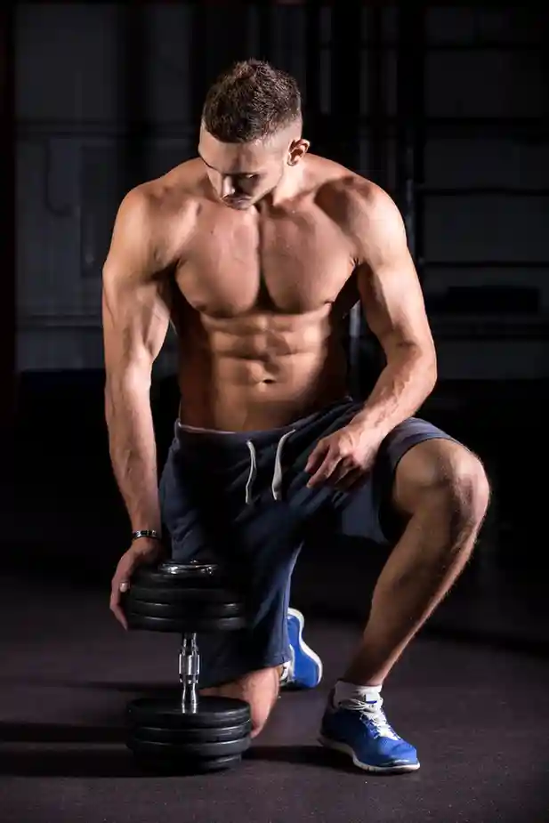 Muscular individual kneeling on one knee in a dimly lit gym, holding a dumbbell in one hand. Wearing blue athletic shorts and matching shoes, the person's defined arms, shoulders, and abs are prominently highlighted against the dark background, emphasizing strength and fitness.<br />
