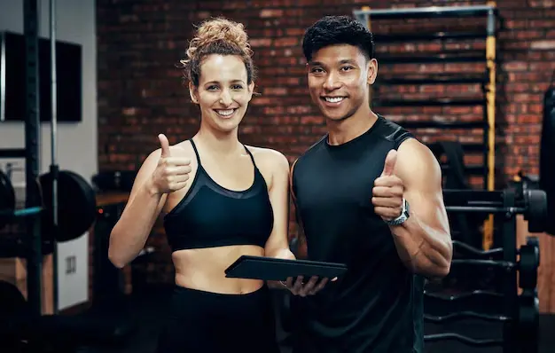 Two individuals in athletic wear pose in a gym, smiling and giving thumbs-up gestures while holding a tablet. The person on the left wears a black sports bra, and the person on the right wears a black sleeveless shirt. Behind them are gym weights and a brick wall, suggesting a fitness or training environment.<br />
