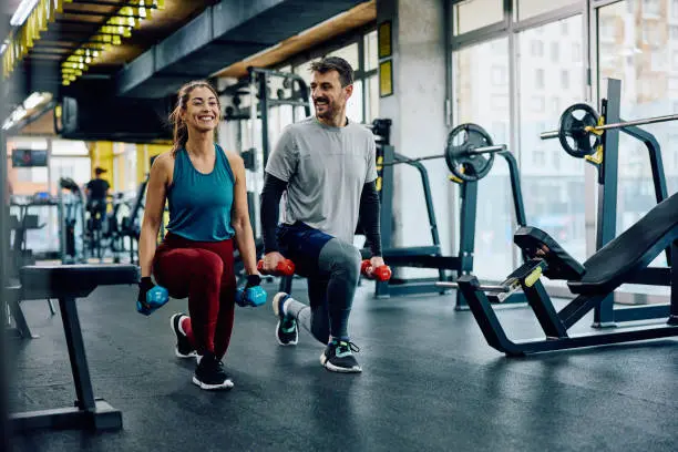 Two individuals performing lunges with dumbbells in a well-equipped gym. One wears a black tank top and gray leggings, the other wears a red T-shirt and black shorts. Both hold dumbbells in each hand, focused on their workout. The gym features weight racks, benches, and large windows with natural light and a city view.