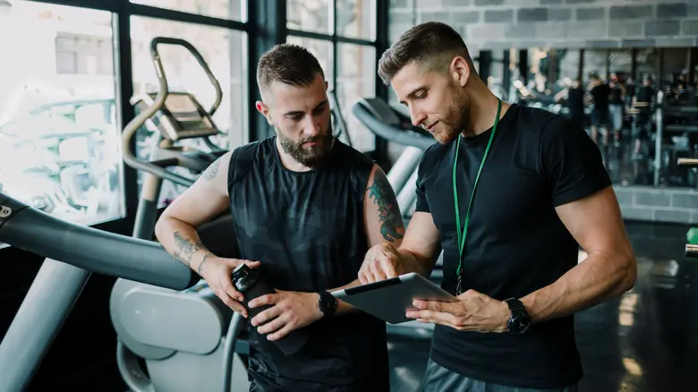 Two individuals in a modern gym setting engage in a focused conversation. One person, holding a water bottle, listens attentively while the other, holding a tablet, appears to explain or demonstrate something. Cardio equipment like treadmills is visible in the background, suggesting a personal training session or fitness consultation.
