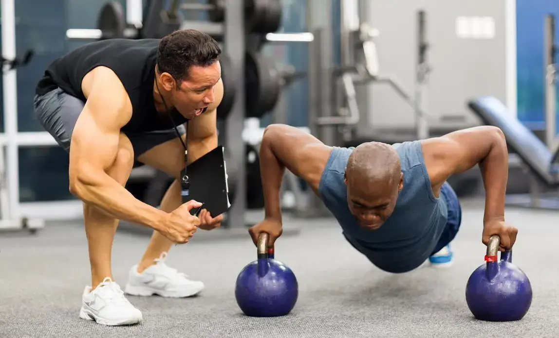 Inside a well-equipped gym, a man in a blue sleeveless shirt performs push-ups using two kettlebells, engaging core and upper body strength. Nearby, a trainer in a black sleeveless shirt and gray shorts observes closely, offering guidance and support. Weight racks and benches fill the background, emphasizing a focused training environment.