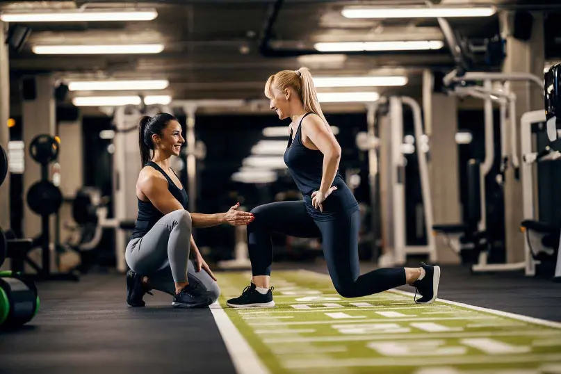 In a gym setting, one individual performs a lunge on a green mat with white markings, focusing on proper form. Another person kneels beside them, offering guidance and support. The background features various exercise machines and weights, emphasizing a personal training session centered on technique and fitness.<br />
