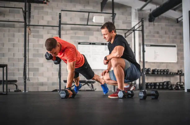 A person performs a plank row exercise in a gym, balancing on one arm while gripping a dumbbell in the other. A coach crouches nearby, observing their form. The gym features concrete walls, a rack of dumbbells, and a whiteboard with notes, creating a focused training environment.
