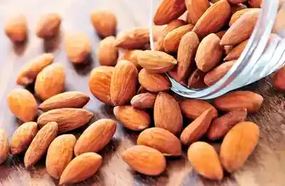 Almonds-MAnsoura City Gym A close-up, elevated view of raw, shelled almonds spilled from a clear glass or jar onto a light wooden surface