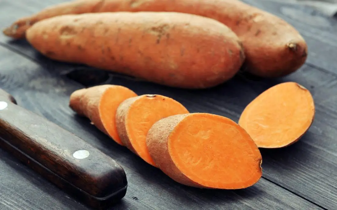 SWEET POTATO-MUSCLE GAINING FOOD-MANSOURA CITY GYM A close-up shot of several raw, whole sweet potatoes and one sweet potato sliced into thick, bright orange rounds, resting next to a knife with a dark wooden handle on a rustic wooden surface.