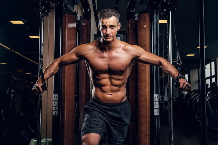 A muscular man with a prominent six-pack and defined arms is performing a cable crossover exercise. He is wearing black gym shorts and has a serious expression, looking directly at the camera. He is standing between two wooden-panelled cable towers in a gym with blurred weights and machines in the background.