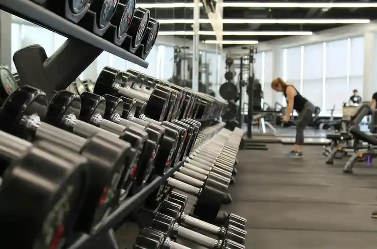 Interior of a modern gym with a neatly organized dumbbell rack in the foreground, featuring metallic weights with black handles arranged by size. The space has rubber flooring, large mirrors, and natural light streaming through tall windows. In the background, a person bends over near benches and weight machines, preparing for a workout.