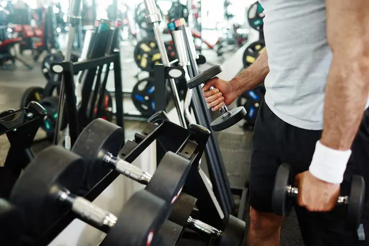 A person in a white t-shirt and black shorts is picking up dumbbells from a rack in a gym.