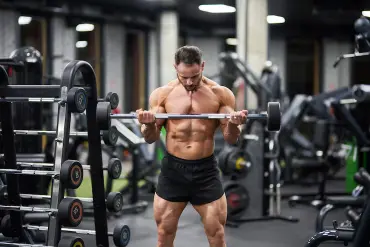 A muscular man is performing bicep curls with a barbell in a gym wearing black shorts, with various gym equipment visible in the background.