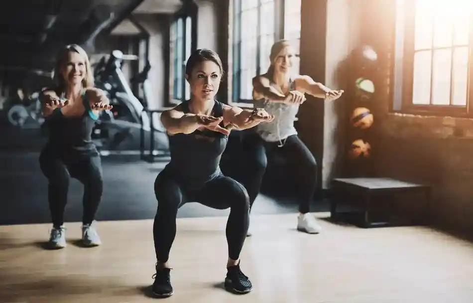 Three ladies performing squats in a gym, aligned in a row with arms extended forward. They are dressed in athletic wear—leggings and tank tops—and appear focused during a group workout session. The background includes stationary bikes, weight machines, and stacked medicine balls near a sunlit window, creating a dynamic and energetic fitness environment.