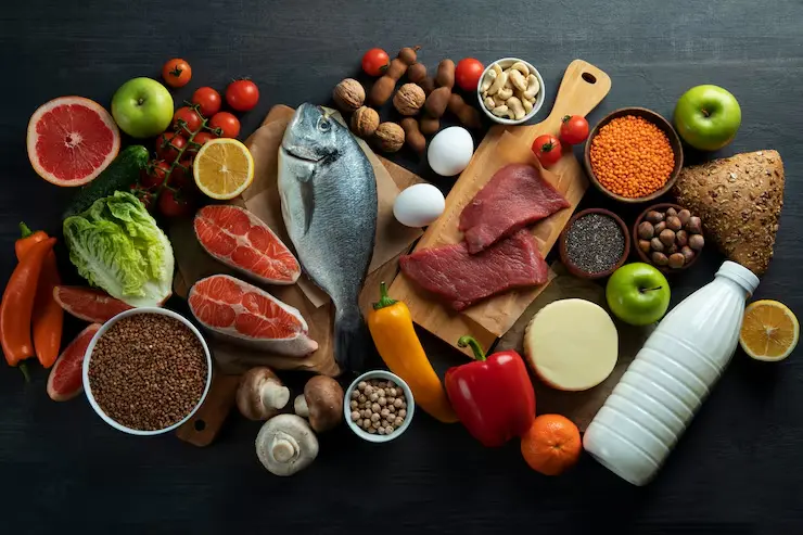 muscle gaining food mansoura city gym A flat lay photo of a variety of whole, unprocessed muscle-gaining foods, including a whole fish, salmon fillets, raw red meat, eggs, a bottle of milk, chickpeas, lentils, walnuts, and a selection of fresh produce like apples, grapefruit, tomatoes, mushrooms, and bell peppers, all arranged on a dark wooden surface.