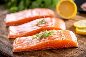 salmon fish-mansoura city gym..muscle gaining foods Three raw salmon fillets, richly orange in color, are arranged diagonally on a wooden cutting board, each garnished with a sprig of fresh dill. A cut lemon and lemon slices are visible in the blurred background.