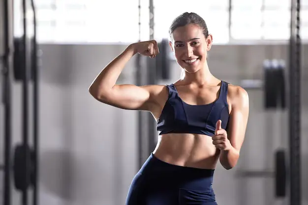 A smiling woman in athletic wear flexes her bicep and gives a thumbs-up in a gym setting.