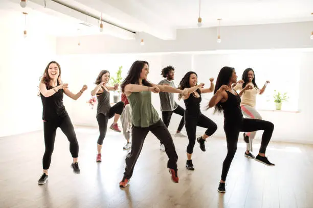 A group of nine people in athletic wear participating in a dance or fitness class in a bright, spacious studio with wooden floors and large windows. They are performing synchronized movements, surrounded by natural light and potted plants near the windows, creating an energetic and lively atmosphere.