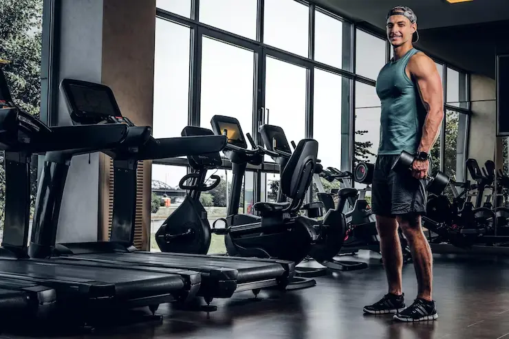 <br />
A color photo of a muscular man smiling while standing in a gym. He is wearing a light blue tank top, dark athletic shorts, a cap, and a pair of headphones. He is holding a dumbbell at his side. The background shows a row of treadmills and exercise bikes positioned in front of large floor-to-ceiling windows that look out onto a tree-lined area. The gym floor is dark and reflective.