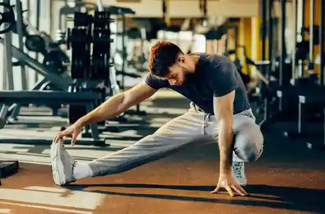 A focused, bearded man wearing a dark t-shirt and grey sweatpants performs a deep lower-body stretch near weight benches in a fitness center.